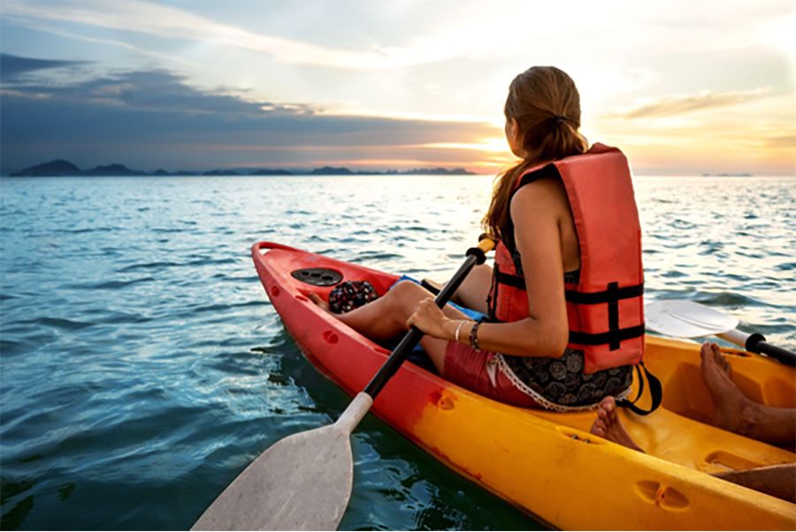 Woman with back to camera wearing lifejacket in an orange and yellow kayak. Paddling out to sea towards sunset.